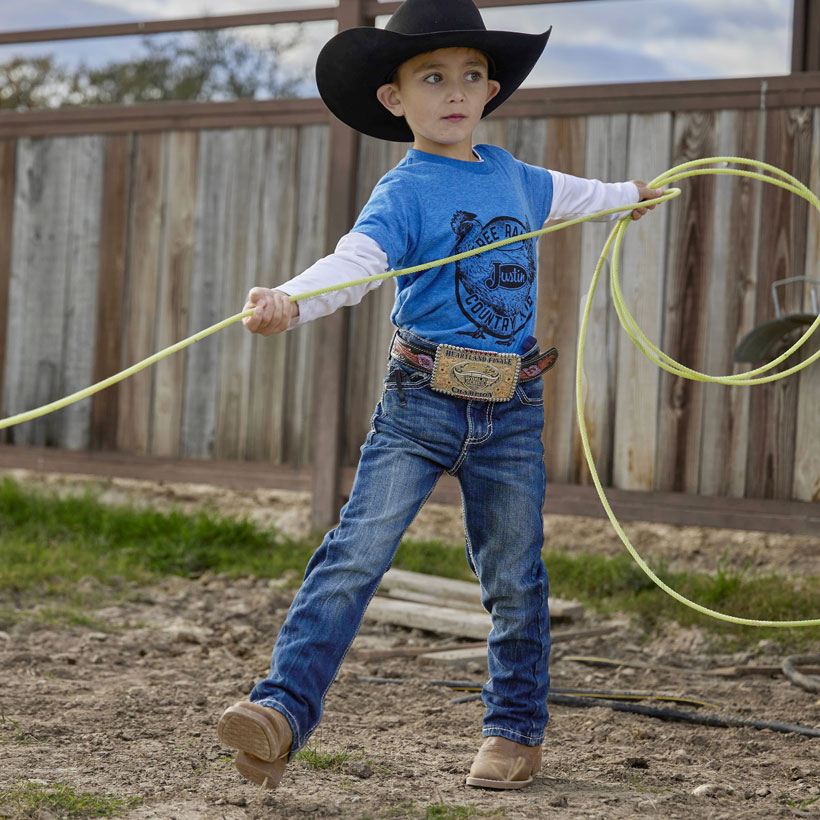 Young kid wearing the Canter Junior boots in Clay wearing jeans, a blue tshirts, and a cowboy hat standing in front of an arena fence roping. 